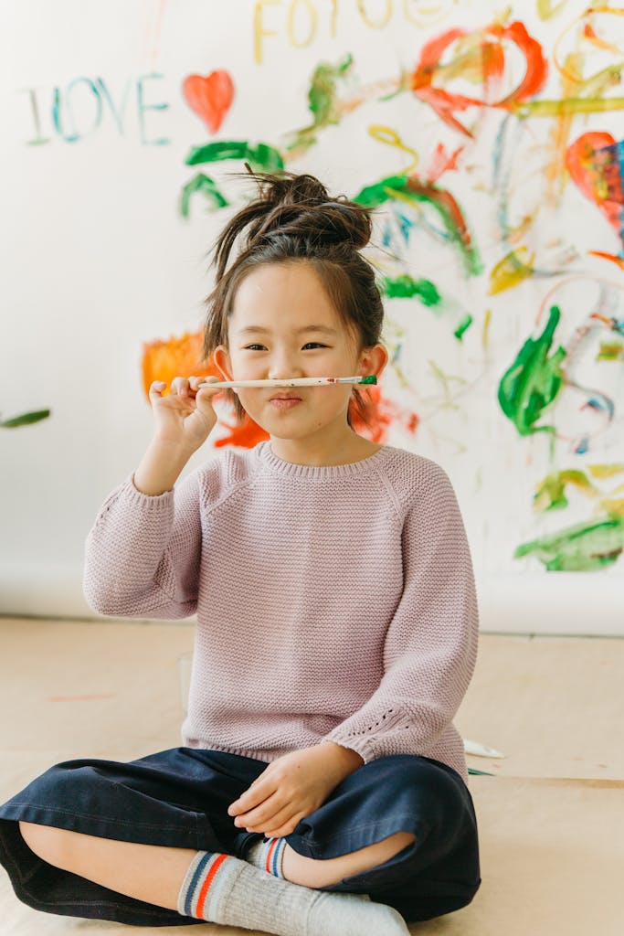Young girl enjoying art by painting with a brush, wearing a pink sweater in a bright setting.