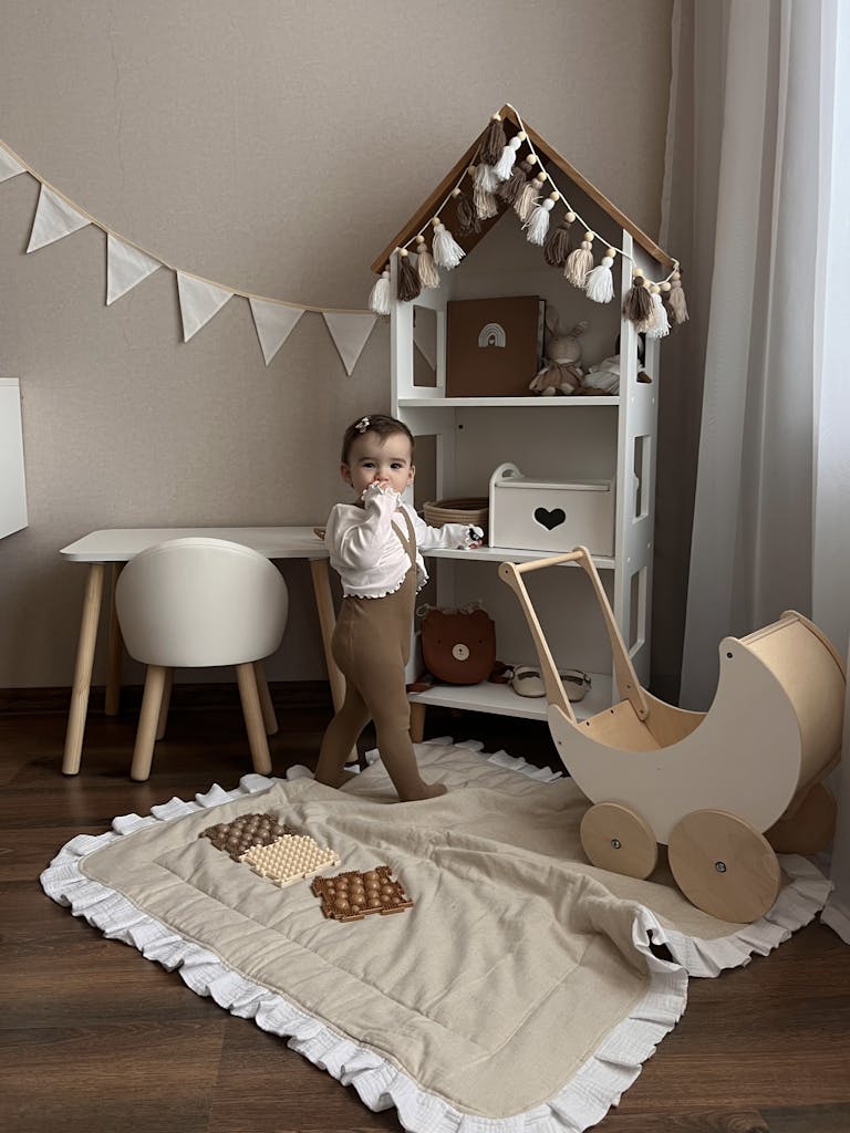 Photo by Карина Суховерхая A baby stands in a stylish playroom with a wooden stroller and toys, evoking warmth.