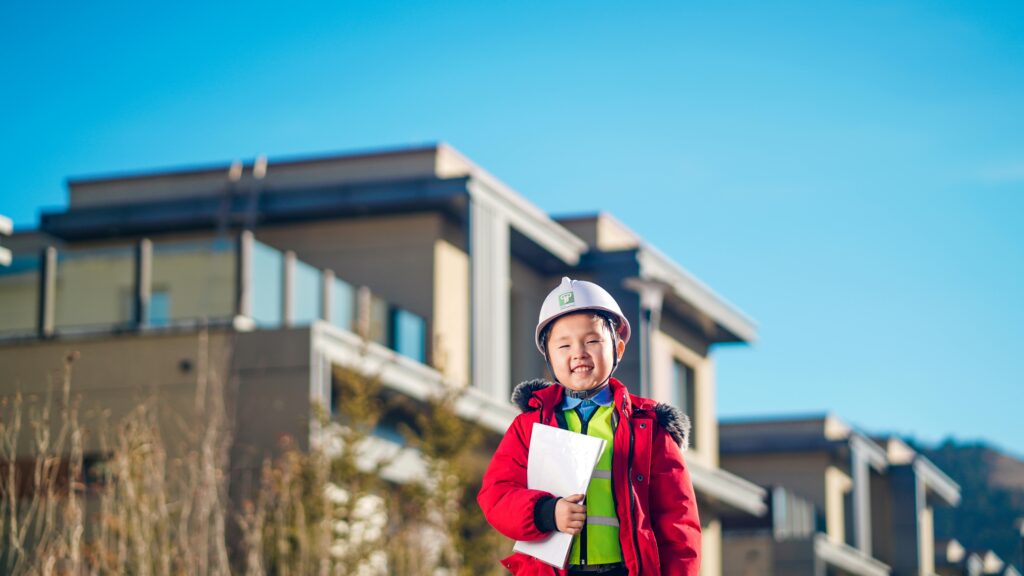 Young child in a hard hat and red jacket smiling in front of buildings.